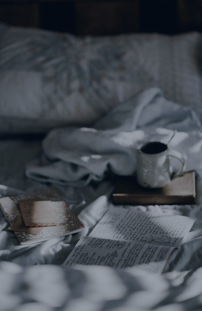 An open handwritten journal on an unmade bed, with a mug of coffee and a notebook nearby