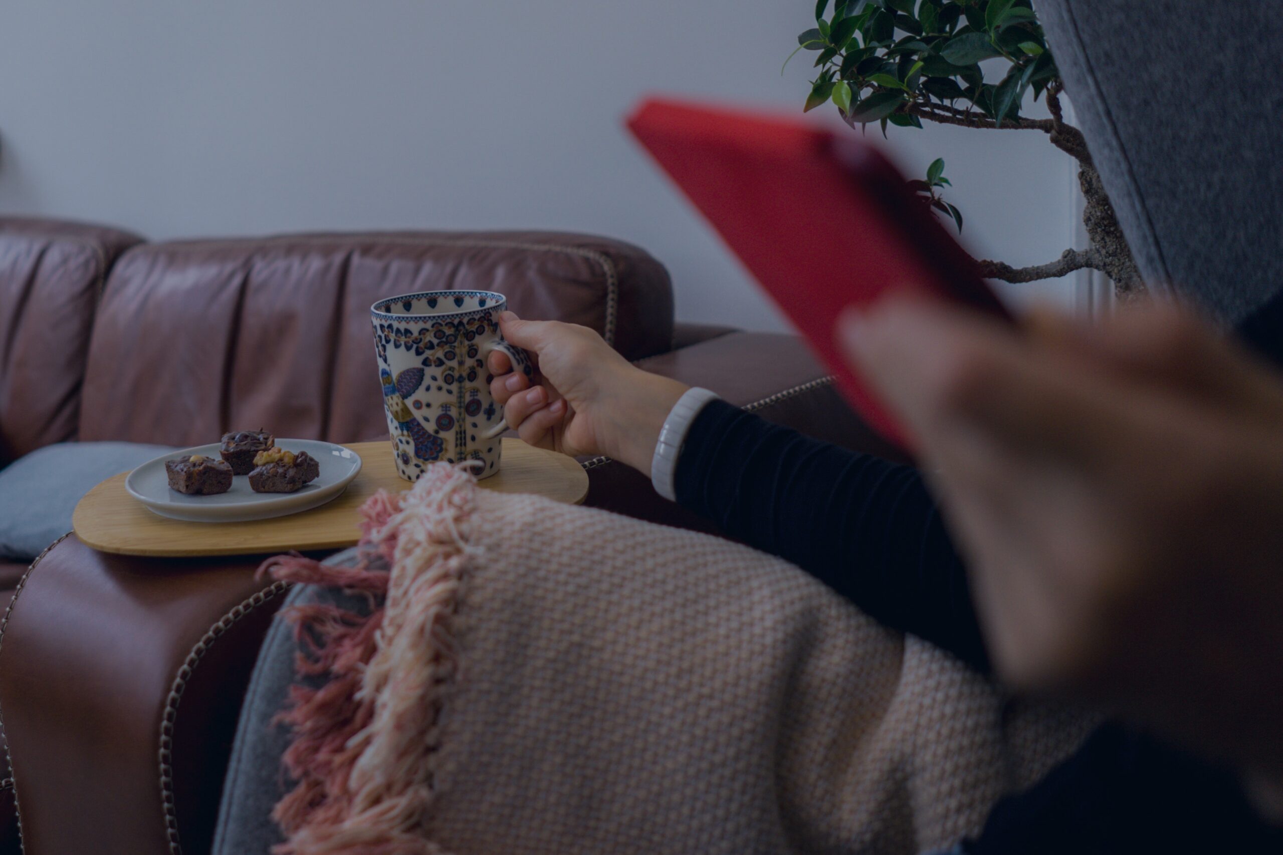A person wrapped in a blanket on a couch, holding a mug of coffee while reaching for a tablet