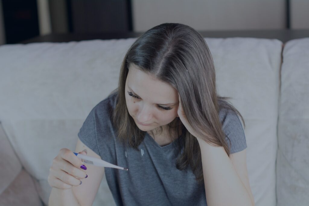 A woman checks her own temperature at home, head resting in her hand — one of the few moments illness gets acknowledged before the day carries on regardless.