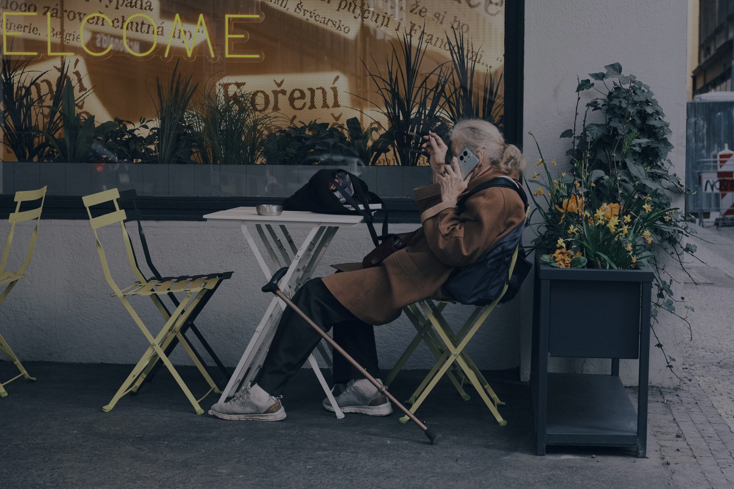An older woman sitting alone at an outdoor café table, on the phone, with an empty chair beside her and a cane resting against the table.